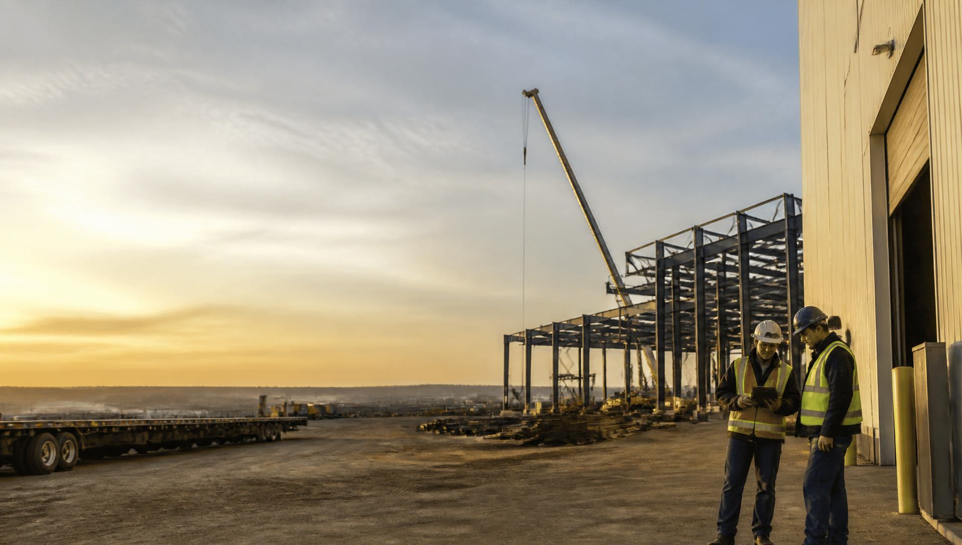 Workers reviewing plans on a tablet beside a steel structure and transport trailers at an Alberta industrial yard at dusk